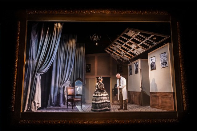 Un ensemble théâtral dans un grand cadre doré, avec une actrice déguisée en Mary Todd Lincoln et un acteur à côté d'elle, portant un manteau blanc