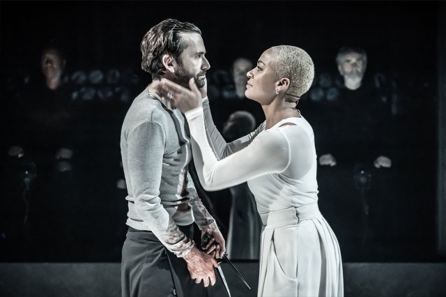An actress grasps the face of an actor on stage, both dressed in monochrome