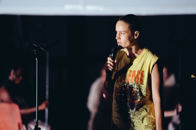 An actress with a shaved head in a yellow sleeveless top holds a microphone on stage