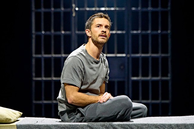 An actor sits on a mattress in a jail cell on stage