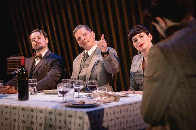 Un groupe d'acteurs dans les costumes d'époque des années 40 est assis à une table à manger sur scène