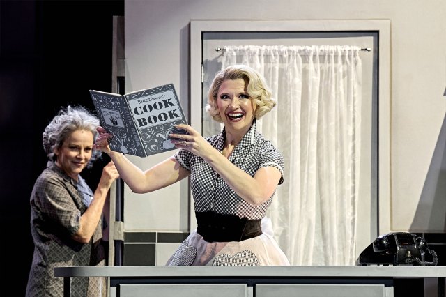 Une actrice sur scène dans un costume de femme au foyer monochrome des années 50, en tenant un livre de cuisine