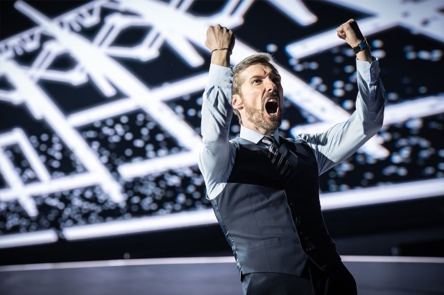 An actor on stage, dressed as football manager Gareth Southgate, raising his arms in celebration in front of a stadium backdrop