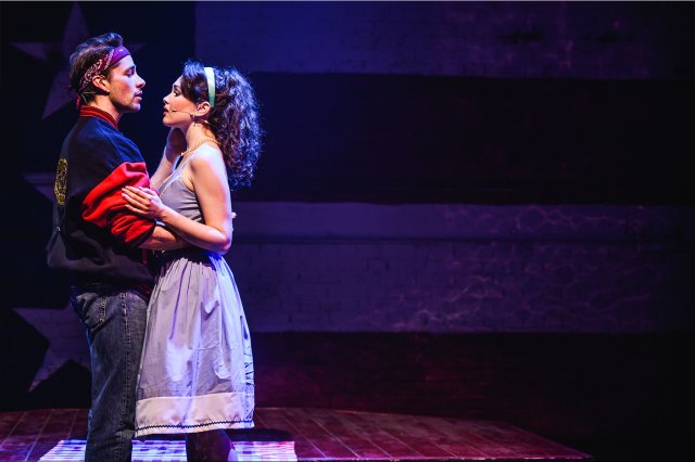 An actor and an actress embrace on stage in front of an American flag-themed backdrop