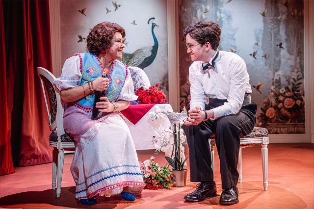 A smartly dressed actress and actor sit on stage in an eloquent room setting