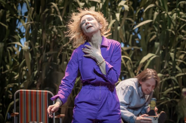 An actress in a purple jumpsuit dances on stage in front of a leafy backdrop
