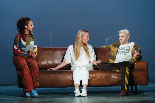 Two actresses and an actor sat on a brown leather sofa on stage