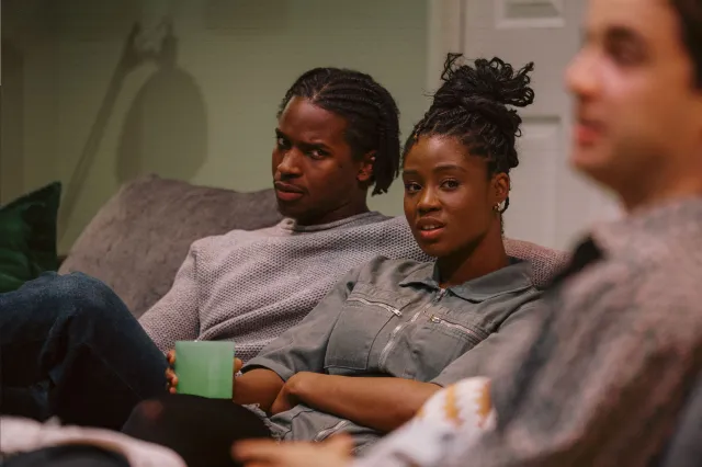 An actor and an actress holding a green mug, sat on a sofa on stage