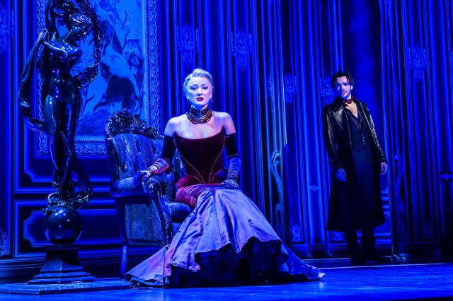 Une actrice dans une belle robe est assise sur scène sur un ensemble luxueux avec un acteur en pardessus en arrière-plan.