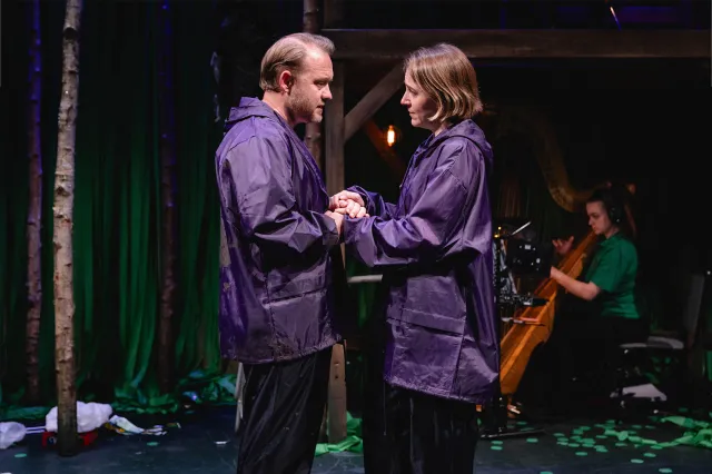 An actor and an actress, both wearing cagoules, hold hands on stage