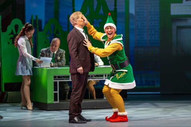 Two actors on stage - one dressed in a business suit and the other in a Christmas elf costume