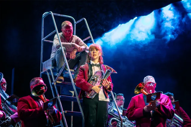 An actress in a brass band uniform, holding a brass instrument, surrounded by band members, with an actor sat on a step ladder behind her.