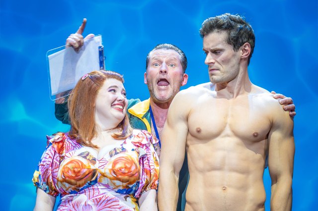 An actress in a flowery dress stands next to a bare-chested actor on stage, with another (clothed) actor behind them, looking over their shoulders.