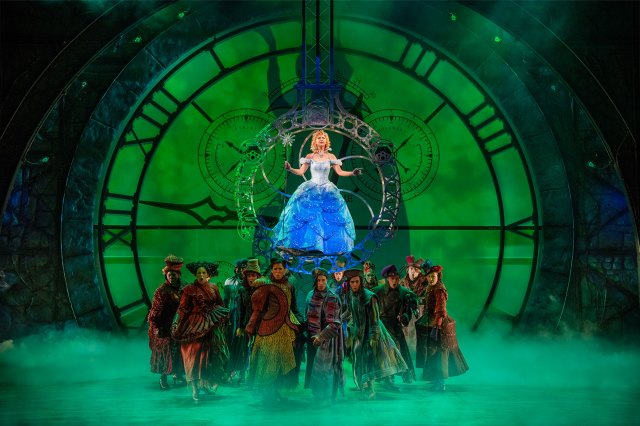 An actress dressed as Glinda the Good hovers above an ensemble cast in front of a huge green clock backdrop.