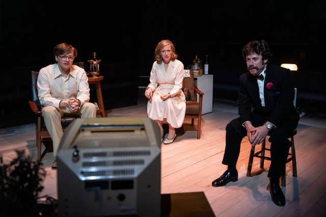 Une actrice avec un acteur de chaque côté d'elle, toutes assises sur scène en regardant un téléviseur.