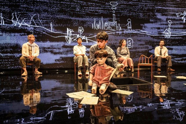 Un acteur avec une marionnette d'un garçon s'agenouille sur le sol de la scène avec plusieurs feuilles de papier, tandis que quatre autres acteurs sont assis sur des chaises en arrière-plan