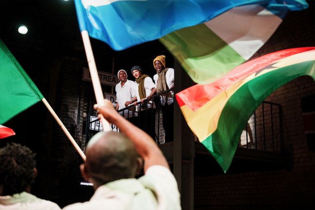 Trois acteurs sur un balcon regardant une foule d'acteurs agitant divers drapeaux des nations africaines.