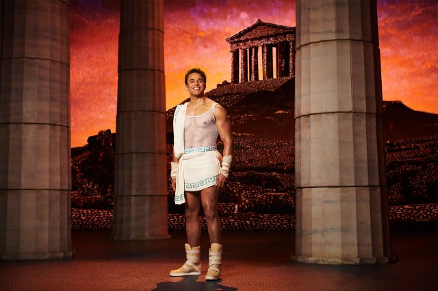 An actor on stage dressed as Hercules, with a Greek temple and columns in the background