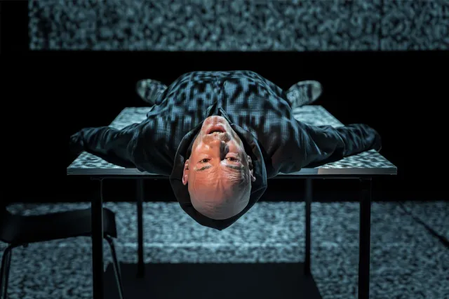 An actor lying on his back on stage with projections of old TV static