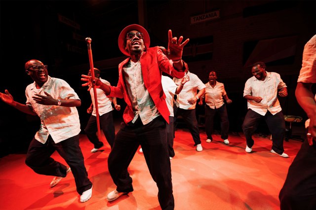A group of African actors on stage, one centered with a red coat, red hat and a cane.