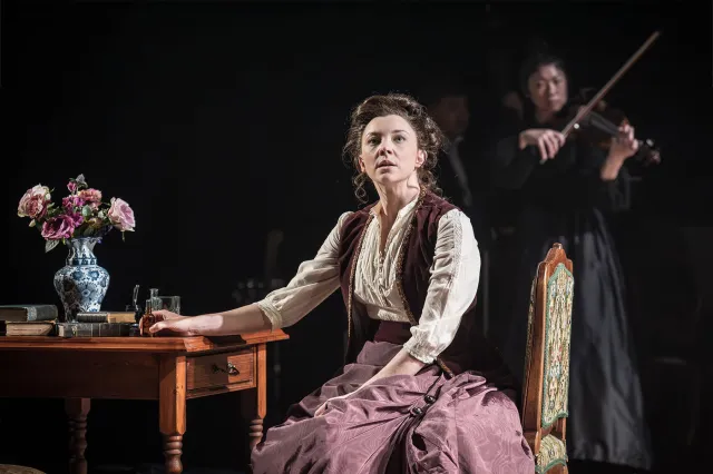 An actress sits at a table on stage, wearing a late 19th-century costume. Another actress plays the violin in the background.