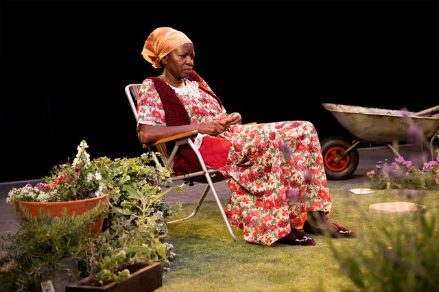 An older actress sits in a garden chair on a stage designed to resemble a garden