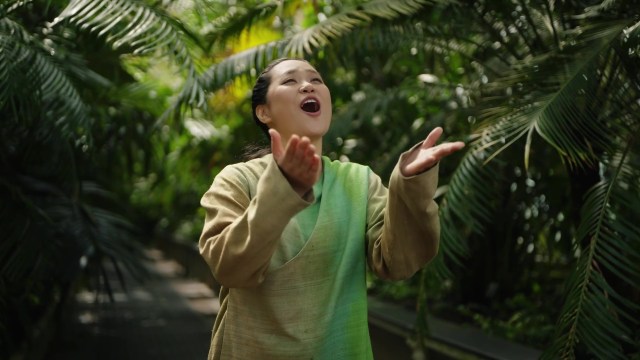 A performer in a green, period Japanese costume performing in a large, plant-filled greenhouse.