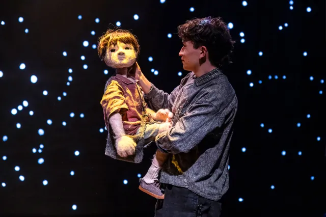 A young actor holding a puppet of a boy, standing in front of a starry backdrop