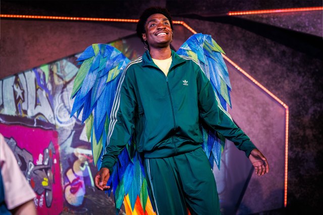An actor on stage, wearing a tracksuit and blue and green wings.