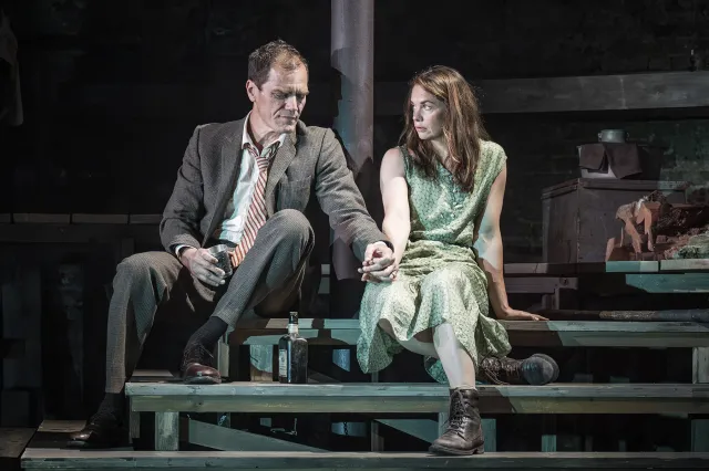 An actor and an actress sit on wooden steps on stage, holding hands.