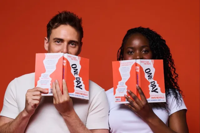 Deux personnes sont assises sur un fond orange, chacune tenant un livre orange vif « ONE DAY » qui couvre une partie de leur visage, portant des hauts blancs.